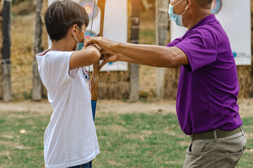 Back view of Asian boy wear face mask aims archery bow and arrow to colorful target in shooting range during training and competition. Exercise and concentration with outdoor archery. Archery for teen