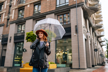 Low-angle view of happy young woman wearing elegant hat standing with transparent umbrella talking on smartphone in rain on European city street, looking away, on background of beautiful building.