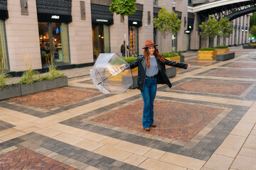 Full length of happy excited young woman in hat dancing and having fun with transparent umbrella on beautiful city street, enjoying rainy weather outdoors. Concept of female lifestyle at autumn season