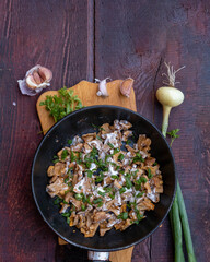 French-fried chanterelle mushrooms with onions and sour cream in a frying pan on a dark wooden background, garlic, onion and parsley and dill greens are laid out next to each other.