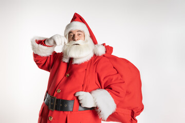 Cheerful Santa Claus with a bag of gifts on his shoulder straightens his mustache while looking at the camera. White background.