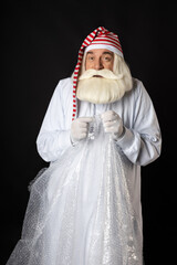 Santa Claus in pajamas and a cap squinted strongly, relaxing, shaking bubble wrap in his hands from excitement. The concept of a cool Santa with a squinted face for Christmas, only Black background.