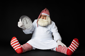 Santa Claus in pajamas sits on the floor with his legs and arms apart, looks at the clock in his...