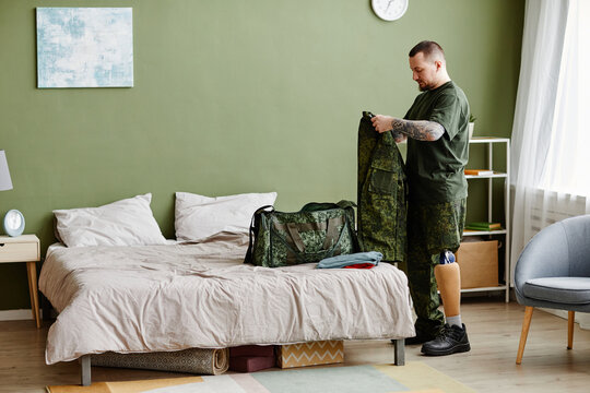 Full Length Portrait Of Military Veteran With Prosthetic Leg Packing Army Uniform At Home, Copy Space