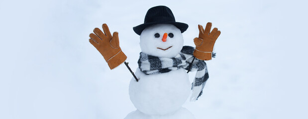 Snowman banner. Funny snowman with a carrot instead of a nose and in a warm hat on a snowy blurred background. Snowman in a scarf, hat and warm winter gloves.