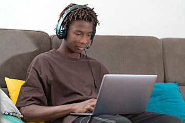 giovane ragazzo nero gamer seduto sul divano comunica con le cuffie mentre usa il laptop  - obrazy, fototapety, plakaty