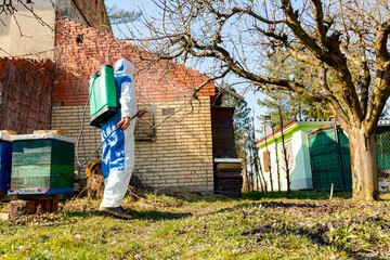 Gardener wearing protective overall sprinkles fruit trees with long sprayer, apiary is in the orchard