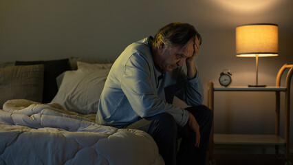Old man holding his head with his hands sits on the bed - Powered by Adobe
