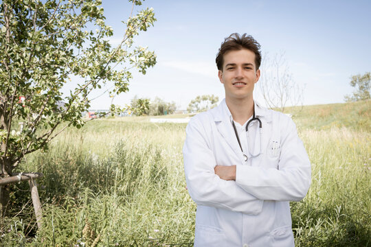 young sympathetic doctor with white coat and stethoscope around his neck stands outside in front of green meadow