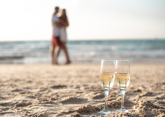 Two glasses with champagne on sand on foreground. Drink wine by seashore. Romantic date of loving...