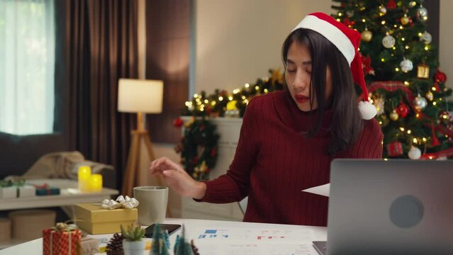 Attractive Asia female business owner wear sweaters sit on table new normal focus online work from house on computer laptop on christmas day relax with drink hot chocolate in mug stay at home working.