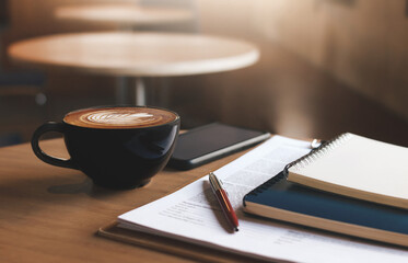 Close up view, latte coffee in black cup with note book and pen on document with mobile phone on...