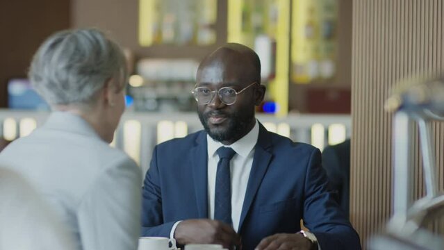 African American businessman greeting Caucasian female colleague with handshake and discussing contract with her while meeting up in restaurant