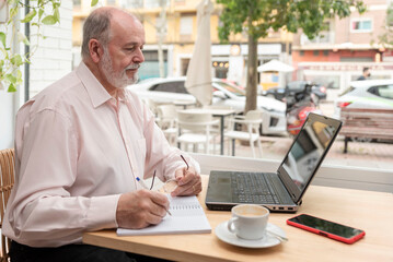 an elderly man sitting at a table with his laptop in front checking the data and taking notes in his notebook, coffee cup and mobile phone next to him