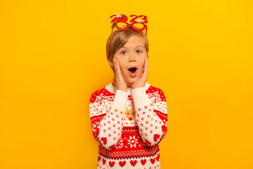 Surprised shocked little kid boy in Christmas winter sweater with party glasses 2023 keeps hands on cheeks and looking at camera on a yellow background.

Happy New Year and Xmas celebration concept.