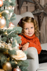 girl in a red sweater sits in an armchair behind a Christmas tree
