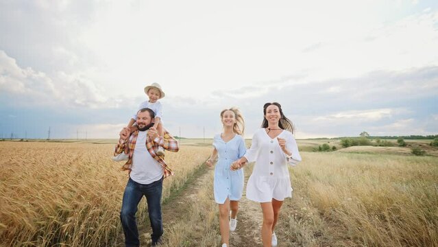 Happy family runs together across summer wheat field with happy toothy smile on their face. Mother and teen girl holding hands. Baby boy sit on shoulders dad and spreading his arms out to sides.