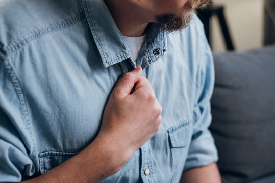 Close Up Sick Man Having Difficulty Breathing Pain Heart, Touches Chest With Hand Sitting On Sofa At Home. Trouble Breathing, Chest Pain. Heart Attack, Thoracic Osteochondrosis, Panic Attack Concept