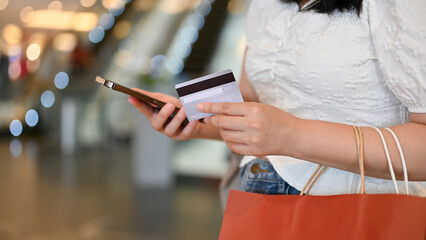 Young Asian woman holding her credit card, using smartphone, walking in the shopping mall