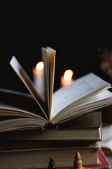 Stack of vintage books, cup of tea or coffee, lit candles, reading glasses and chess pieces on wooden table. Dark academia concept. Selective focus.