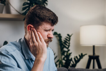 Close-up of upset young guy 30s touches sore ears suffering from sudden throbbing earache....