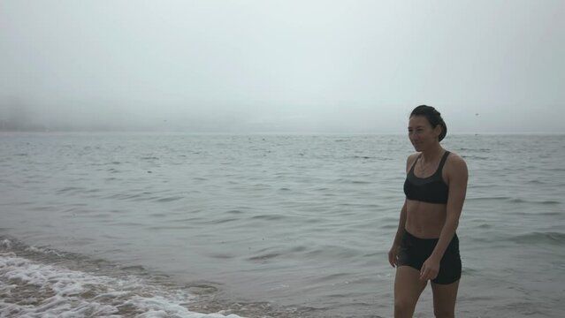 A girl in a black swimsuit comes out of the ocean with waves into the fog, having received pleasure.