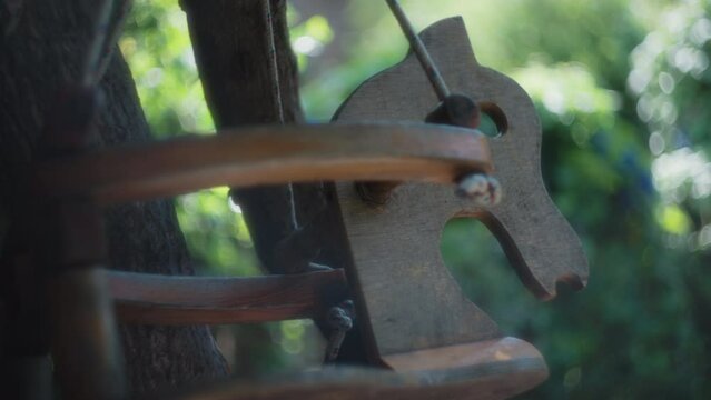 Wooden child swing in a shape of a horse next to a tree trunk. Natural bokeh background, slow motion, shallow depth of field. 