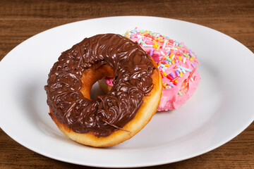 Sweet donuts with chocolate icing and another donuts with pink icing and colorful sprinkles served on a plate. Close-up photography with food centered.