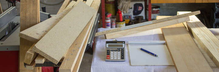 Image of a workbench in a workshop with a calculator, notepad, and wooden boards on it. Reference to the increase in the cost of timber. Horizontal banner 