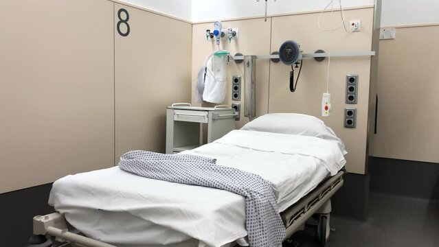 Slowmotion shot of a hospital cubicle with one bed and some medical instruments. White bed with a patient coat on the bed and the alarm button dangling. Hospital patient room with a bed.