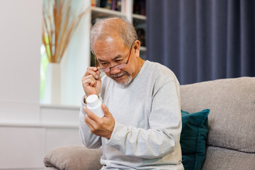 worried senior Asian man taking prescription medicine at home. older man reading drug label with farsightedness problem. - obrazy, fototapety, plakaty