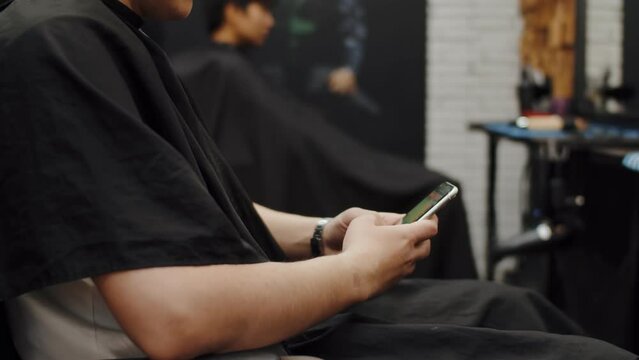 Side view of a man using smartphone in barbershop. Hipster guy sitting in chair holding his smartphone in hands and chatting scrolling in hair salon