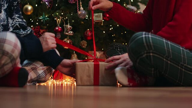 two female hands untie bow on Christmas gift box together. female couple friends sit floor Christmas tree unpacking wrapped craft paper box tied red ribbon bow holding hands present unpack surprise