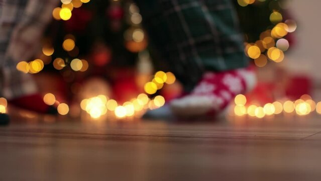 dancing legs in Christmas socks and pajamas dance near Christmas tree golden lights festive garland wooden floor indoors. Merry Christmas and Happy New Year celebration winter holidays concept