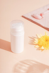 White cosmetic bottle, yellow flower and facial roller on beige background in sunlight. Skincare, spa and wellness concept