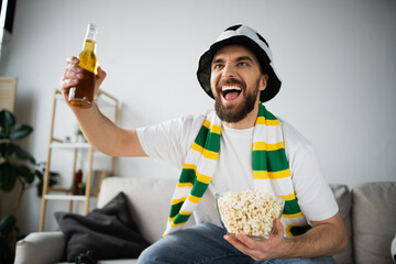 excited sports fan in hat and scarf holding bowl with popcorn and bottle of beer while watching championship. - obrazy, fototapety, plakaty