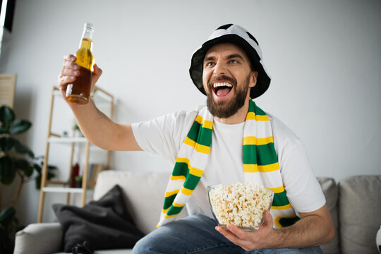 excited sports fan in hat and scarf holding bowl with popcorn and bottle of beer while watching championship.