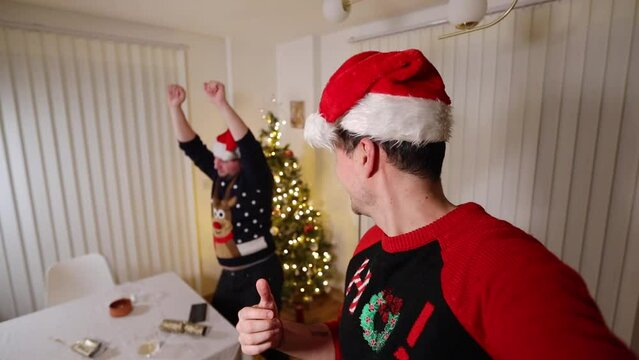Christmas guys wearing festive hats and sweaters vlogging looking at the camera laughing and dancing next to a Christmas tree