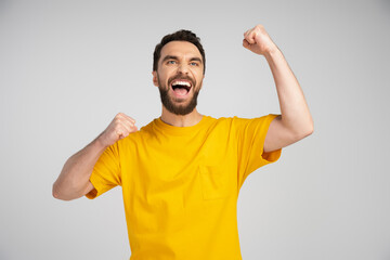 joyful bearded man screaming and showing triumph gesture while looking away isolated on grey.