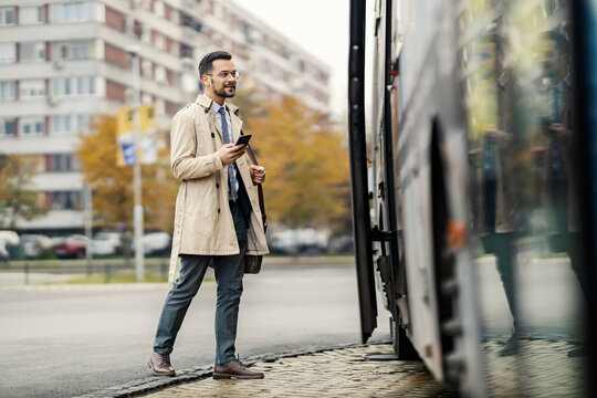 A businessman is using his phone and entering a bus downtown.