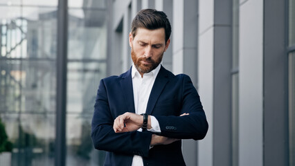 Professional confident businessman standing outdoors waiting for meeting looks at wristwatch checking time displeased caucasian man pointing on watch looking at camera dissatisfied with being late