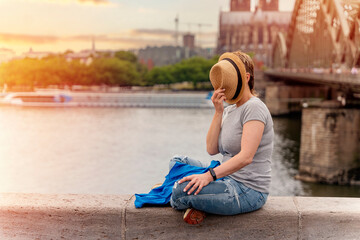 Fototapeta na wymiar woman in hat and blue shirt sitting on embankment of Rhine on background of Cologne Cathedral and Hohenzollern Bridge in Koel, Germany. Tourism and travel by Germany concept