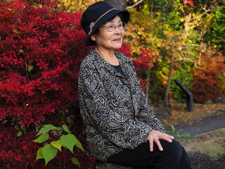 紅葉する木々をバックに自然公園のベンチに座って休憩する高齢日本人女性