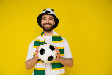 happy man in football fan hat and striped scarf holding soccer ball isolated on yellow.