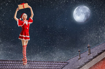 Dancing girl ballerina with a gift in her hands in a Santa Claus costume on the roof of a house on Christmas night against the background of the starry sky.