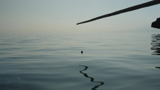 I am fishing in the sea with a homemade bamboo fishing rod, the float is swaying on the waves. Close-up. Mountains in the background.