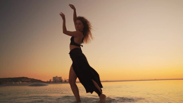 Beautiful girl dancer dancing in water at sunset light. Sensual dance. Barefoot woman dressed black dancing seductive in slow motion, freedom concept. Freestyle contemporary dance Low angle view