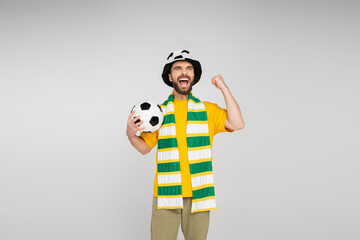 Fototapeta premium excited sports fan in hat and scarf holding soccer ball and showing triumph gesture isolated on grey.