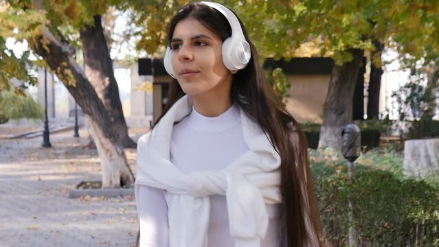 Portrait: a beautiful long-haired Middle Eastern brunette in white clothes and headphones walks in the park listening to her favorite music. Attractive woman enjoying music in the park in autumn