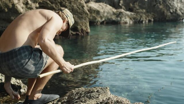 A man in shorts is fishing in the sea with a homemade bamboo fishing rod, he sits on a stone by the water.